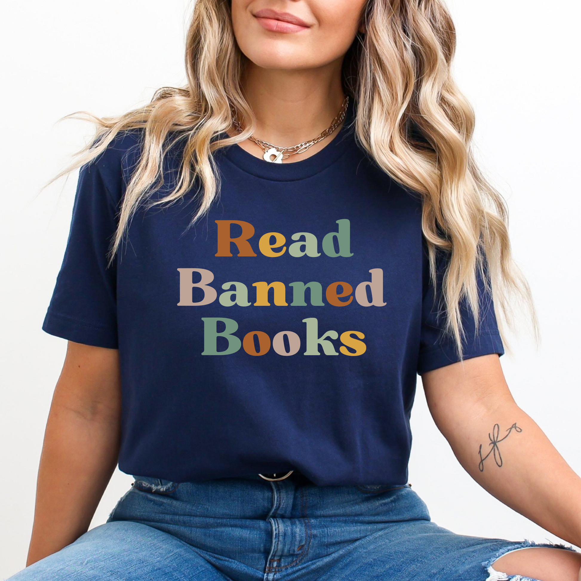 Person wearing a navy blue t-shirt with 'Read Banned Books' text in colorful letters. read banned books shirt, banned books tee, book lover shirt, librarian shirt, teacher book shirt, freedom to read shirt, literary shirt, bookish apparel, reading shirt, library activism shirt, anti censorship shirt, intellectual freedom tee, book club shirt, educational shirt, reader gift shirt
