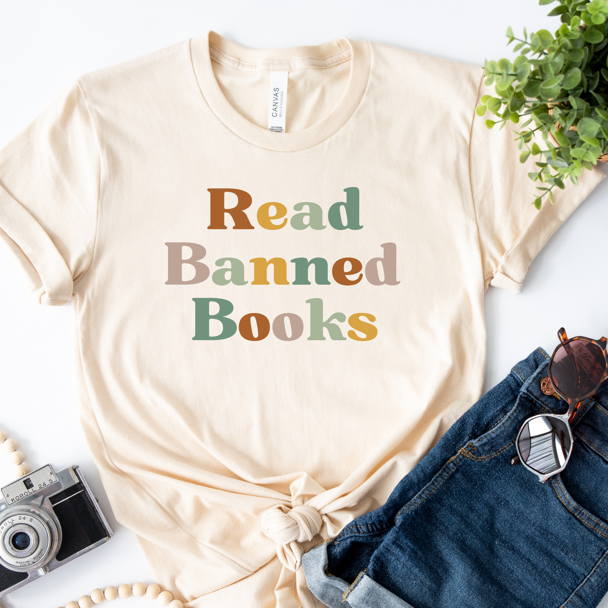 Beige t-shirt with 'Read Banned Books' text on a white surface with jeans, camera, and plant. read banned books shirt, banned books tee, book lover shirt, librarian shirt, teacher book shirt, freedom to read shirt, literary shirt, bookish apparel, reading shirt, library activism shirt, anti censorship shirt, intellectual freedom tee, book club shirt, educational shirt, reader gift shirt
