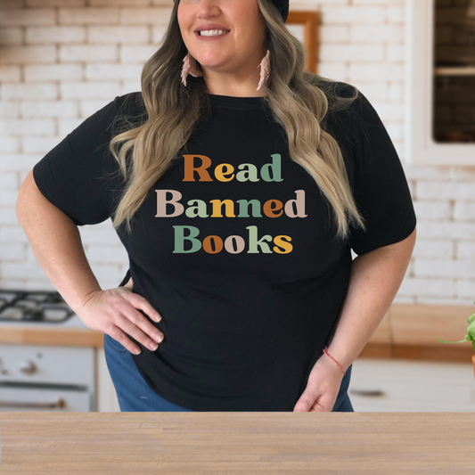 Person wearing a black t-shirt with 'Read Banned Books' text in colorful letters in a kitchen setting. read banned books shirt, banned books tee, book lover shirt, librarian shirt, teacher book shirt, freedom to read shirt, literary shirt, bookish apparel, reading shirt, library activism shirt, anti censorship shirt, intellectual freedom tee, book club shirt, educational shirt, reader gift shirt