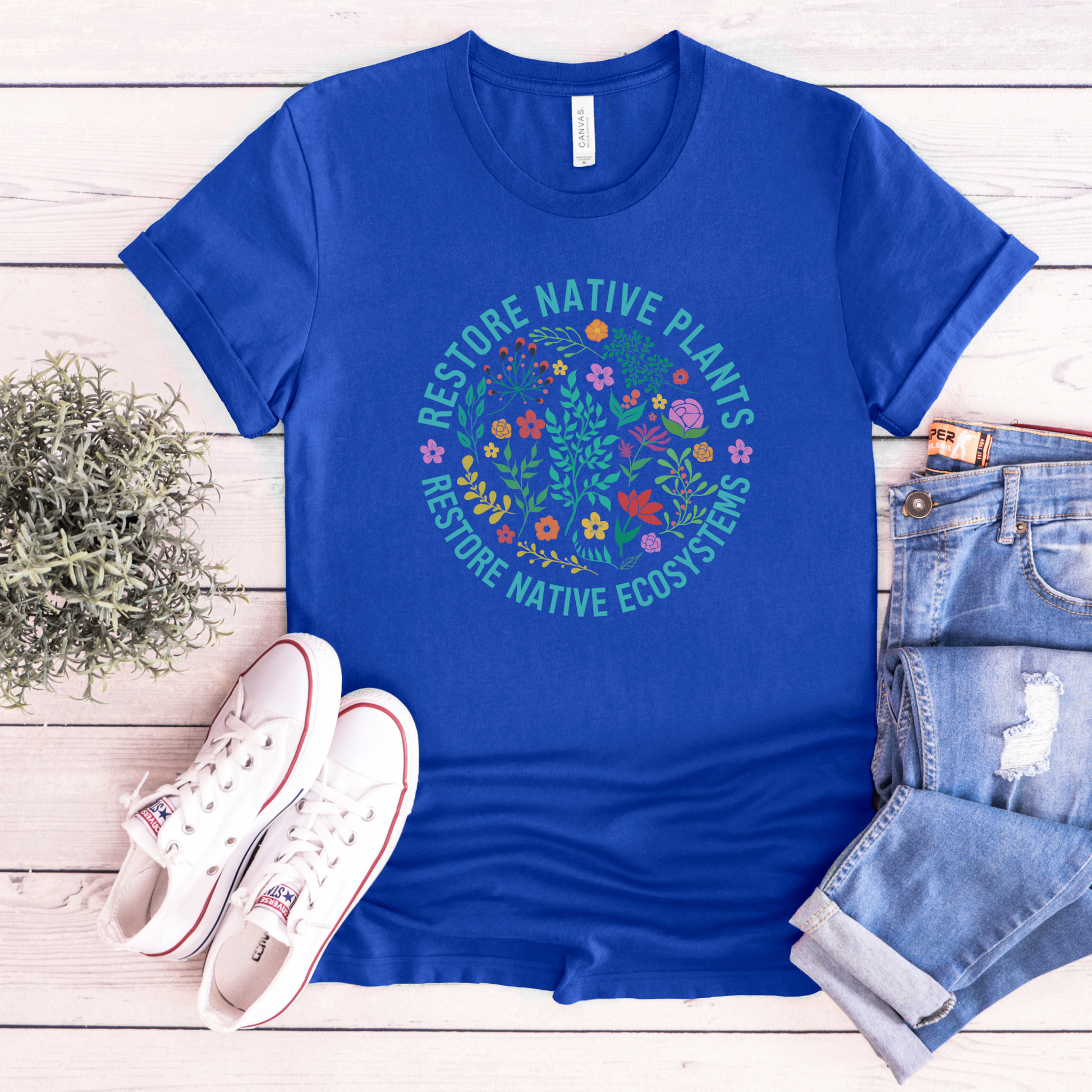 Blue t-shirt with nature-themed design on a wooden surface with shoes and jeans. restore native plants shirt, native plants tee, indigenous plant shirt, land restoration shirt, plant lover tee, environmental advocacy shirt, native plant lover shirt, nature graphic tee, unisex plant shirt, eco friendly apparel, indigenous inspired shirt, stewardship t shirt
