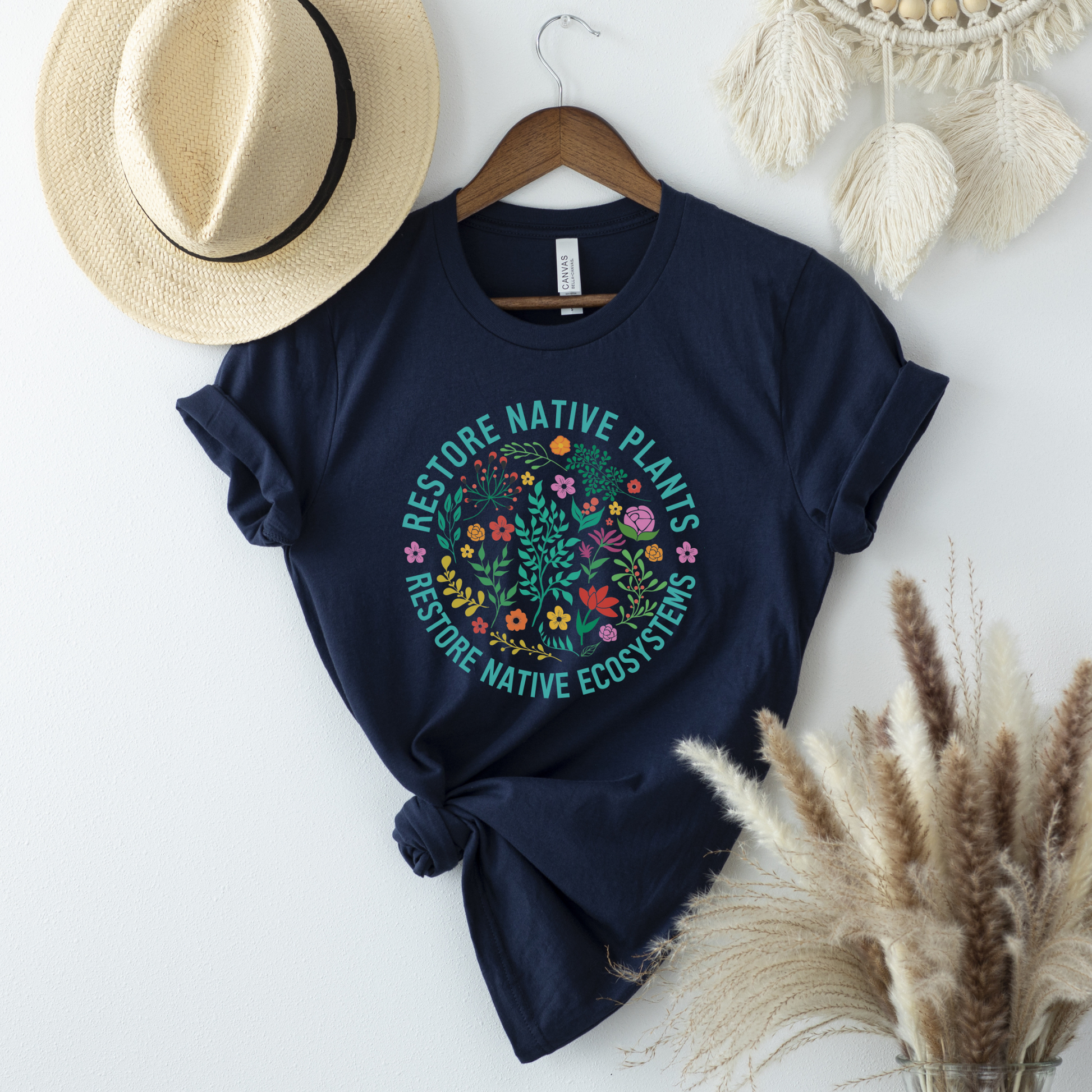 Navy blue t-shirt with nature-themed design on a white surface with decorative items. restore native plants shirt, native plants tee, indigenous plant shirt, land restoration shirt, plant lover tee, environmental advocacy shirt, native plant lover shirt, nature graphic tee, unisex plant shirt, eco friendly apparel, indigenous inspired shirt, stewardship t shirt