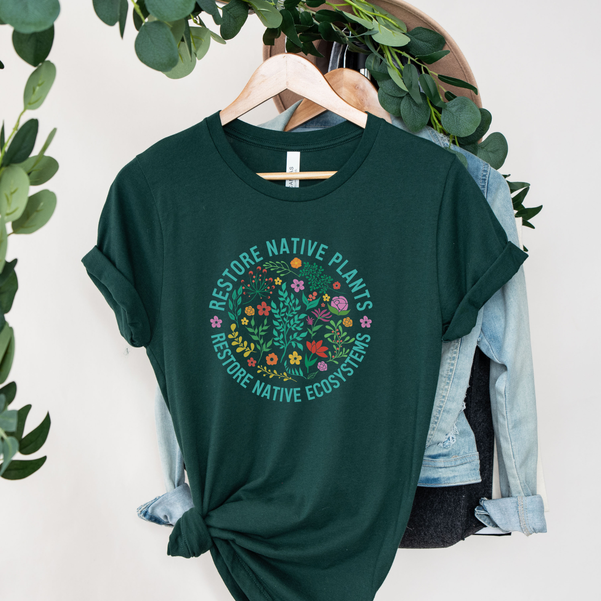 Green t-shirt with 'Restore Native Plants' design on a hanger with a light blue jacket and greenery restore native plants shirt, native plants tee, indigenous plant shirt, land restoration shirt, plant lover tee, environmental advocacy shirt, native plant lover shirt, nature graphic tee, unisex plant shirt, eco friendly apparel, indigenous inspired shirt, stewardship t shirt