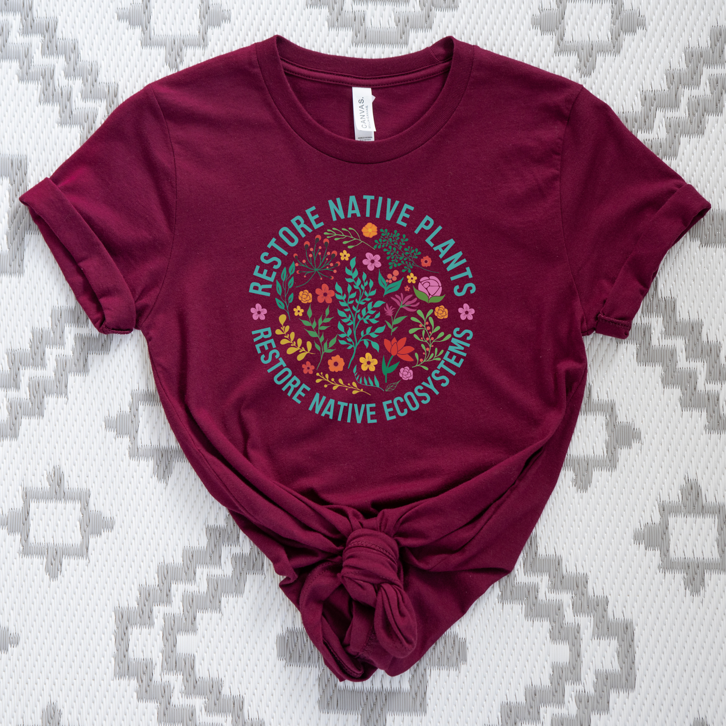 Maroon t-shirt with nature-themed design on a patterned surface. restore native plants shirt, native plants tee, indigenous plant shirt, land restoration shirt, plant lover tee, environmental advocacy shirt, native plant lover shirt, nature graphic tee, unisex plant shirt, eco friendly apparel, indigenous inspired shirt, stewardship t shirt