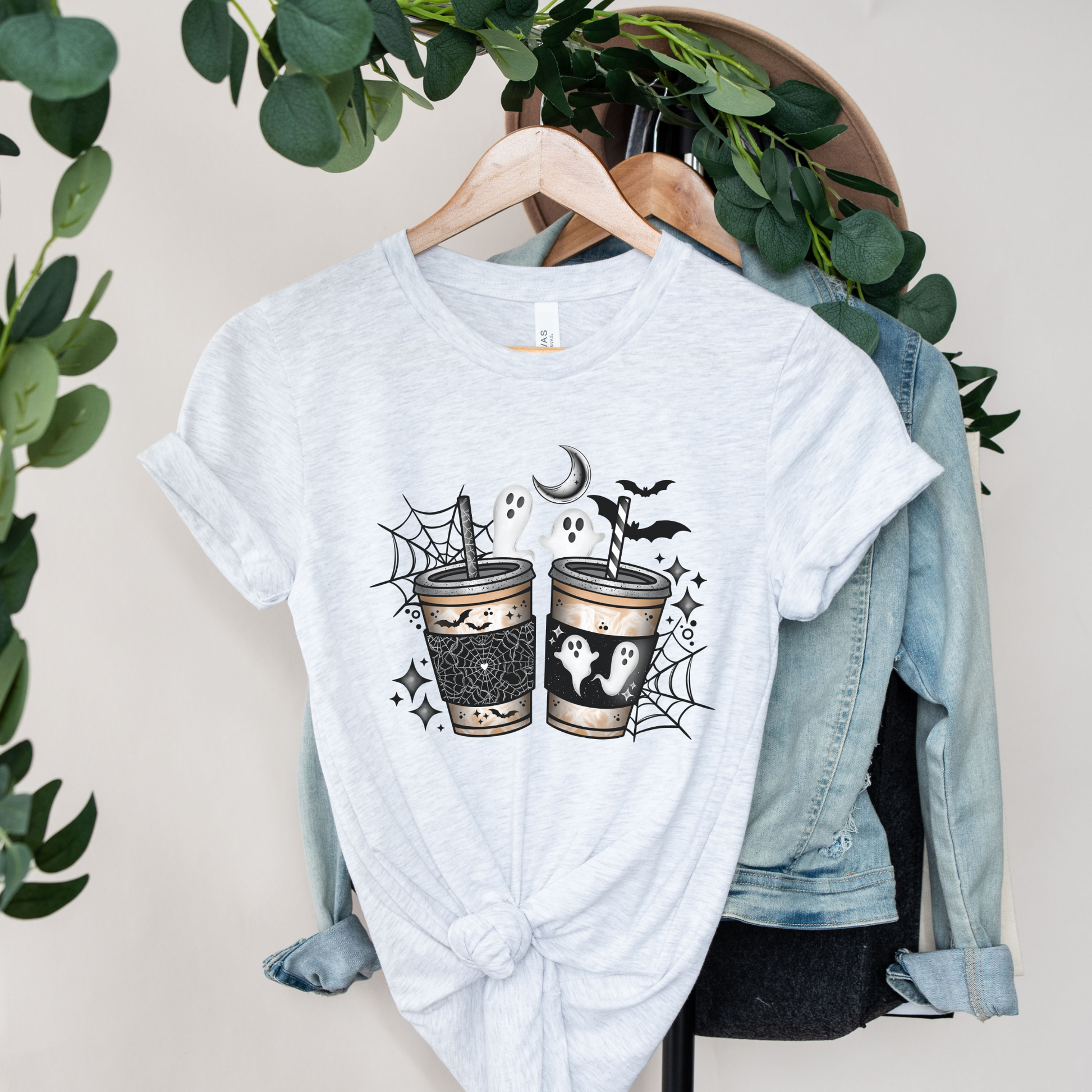 A light heather gray t-shirt with a Halloween-themed graphic print of two decorated cups, a ghost, and bats, styled on a wooden hanger with a denim jacket and green foliage in the background.