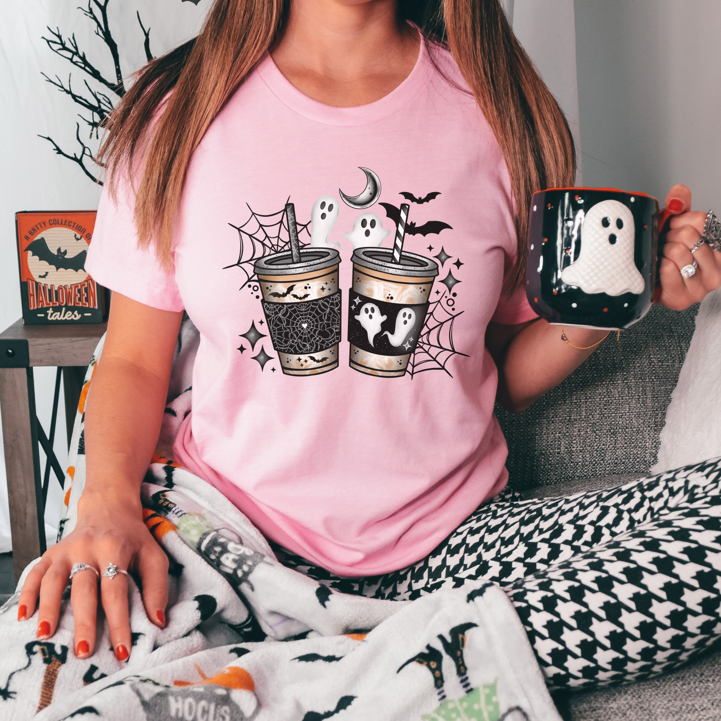 A woman wearing a light pink graphic t-shirt with a Halloween theme, sitting on a couch and holding a black mug with a white ghost design. She is also wearing a black and white houndstooth patterned skirt and a white blanket with black spider graphics.