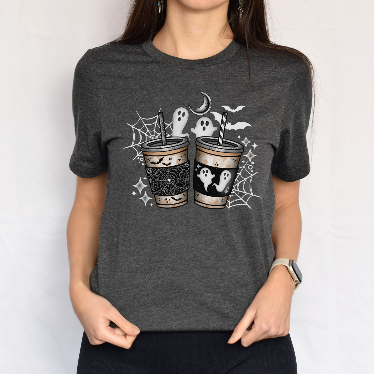 A woman wearing a dark heather gray t-shirt with a Halloween graphic of two coffee cups decorated with ghosts, spiderwebs, and bats. ghost coffee shirt, spooky coffee shirt, halloween coffee tee, cute ghost shirt, spooky season shirt, pumpkin spice shirt, fall coffee lover, halloween graphic tee, ghost aesthetic shirt, coffee lover gift, cozy fall outfit, pumpkin patch shirt, autumn halloween tee