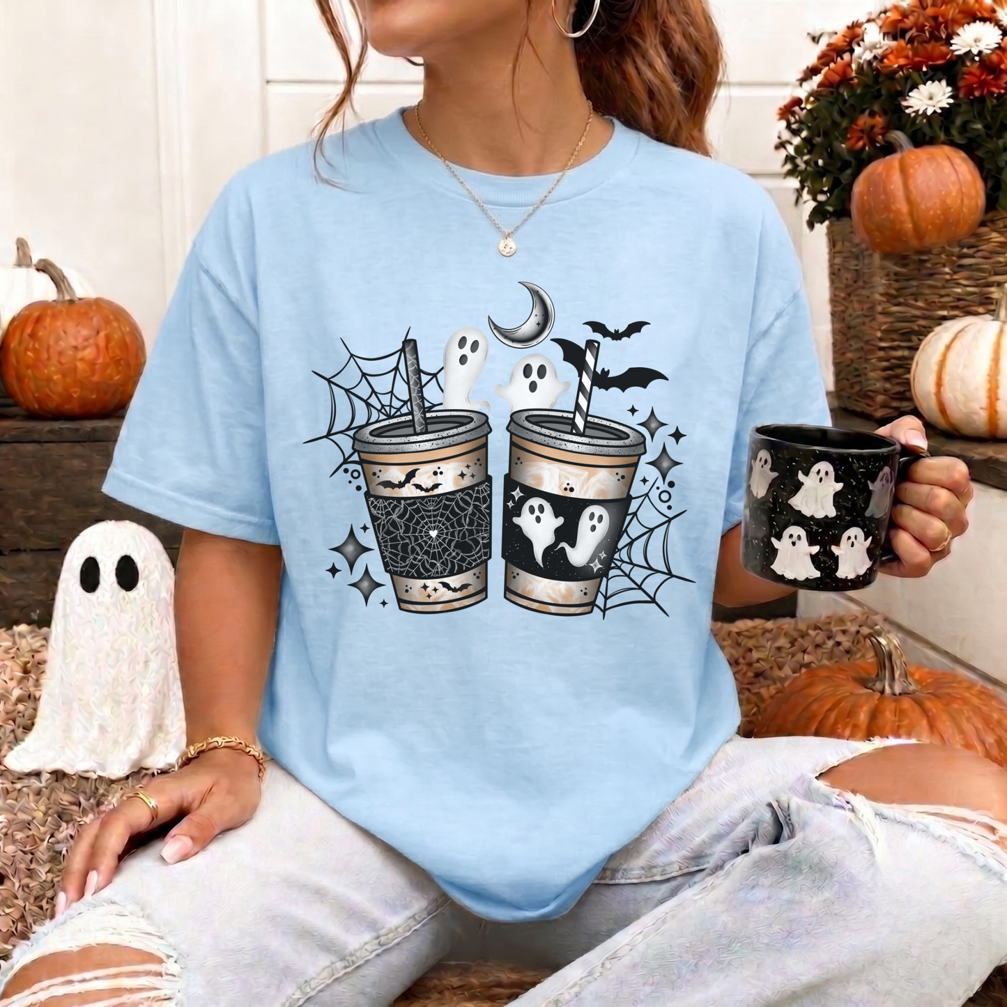 A woman wearing a light blue graphic t-shirt with a Halloween design featuring two cups, ghosts, bats, and a crescent moon. She is holding a black mug with white ghost patterns, surrounded by autumn decorations like pumpkins and a white ghost figurine.