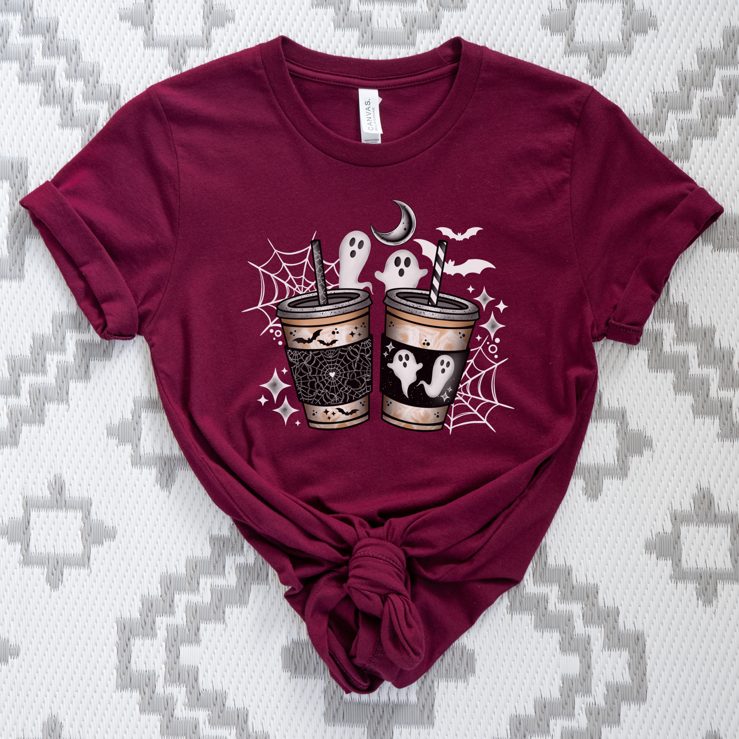 A maroon short-sleeve t-shirt with a Halloween graphic of two laced-up coffee cups, ghosts, bats, a crescent moon, and spiderwebs, styled with a knot at the bottom on a gray and white patterned background.