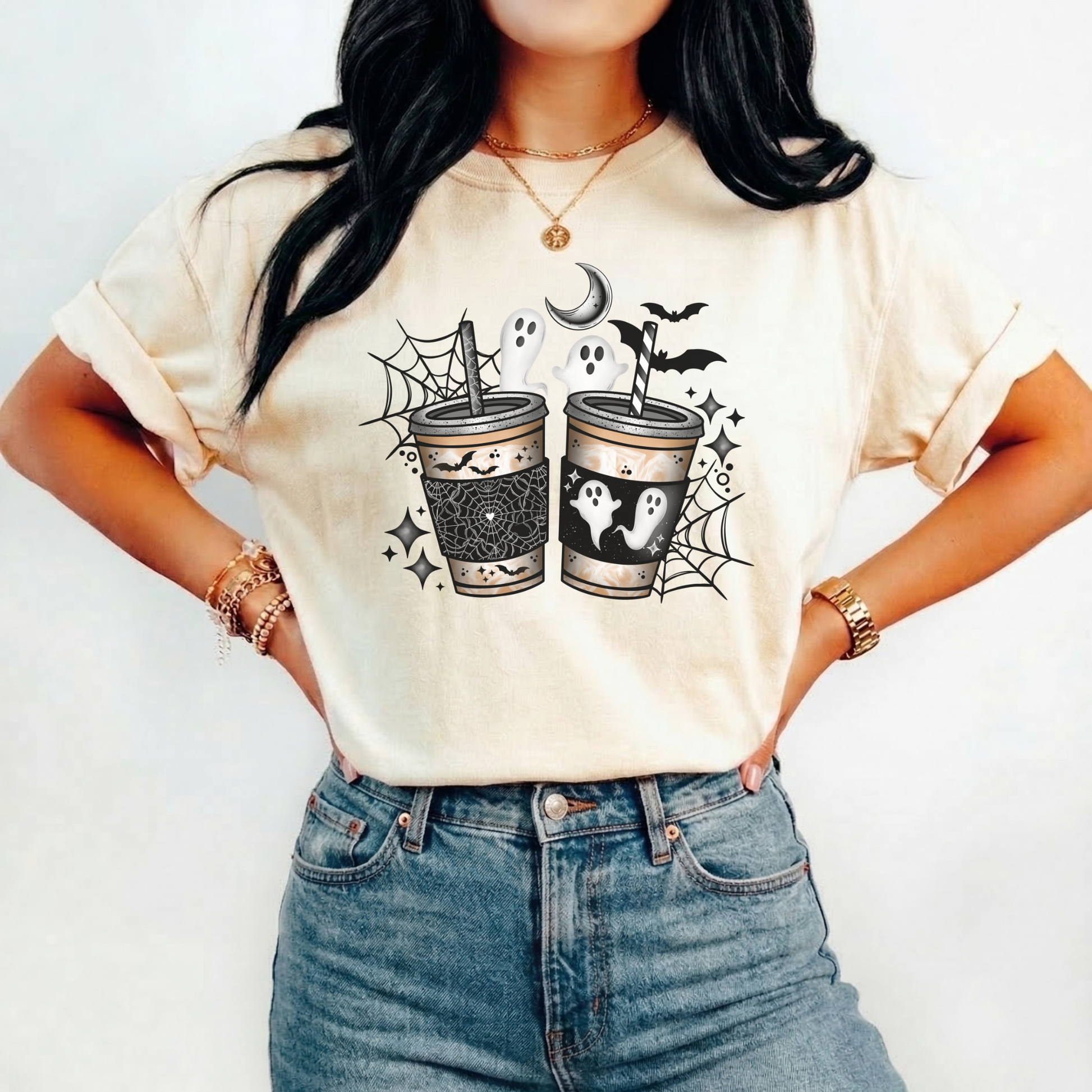 A woman wearing a cream-colored graphic t-shirt with a Halloween-themed design of two coffee cups with ghosts, spiders, and bats, paired with blue jeans.