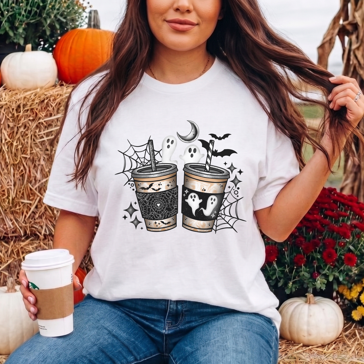 A woman wearing a white graphic t-shirt with a Halloween-themed coffee cup design, sitting in a fall-themed setting with pumpkins and hay.
