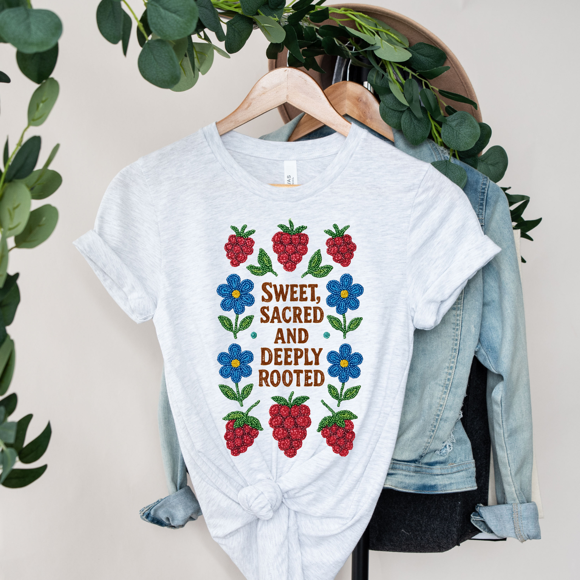 White t-shirt with floral and berry design on a hanger with a denim jacket and greenery. sweet sacred deeply rooted shirt, beaded floral berry tee, indigenous beaded design shirt, sacred roots indigenous apparel, floral berry cultural t-shirt, original indigenous graphic tee, beaded indigenous shirt, deeply rooted heritage tee, unisex cultural pride shirt, sahdeh spruce indigenous design, berry beadwork t-shirt, ancestral connection apparel