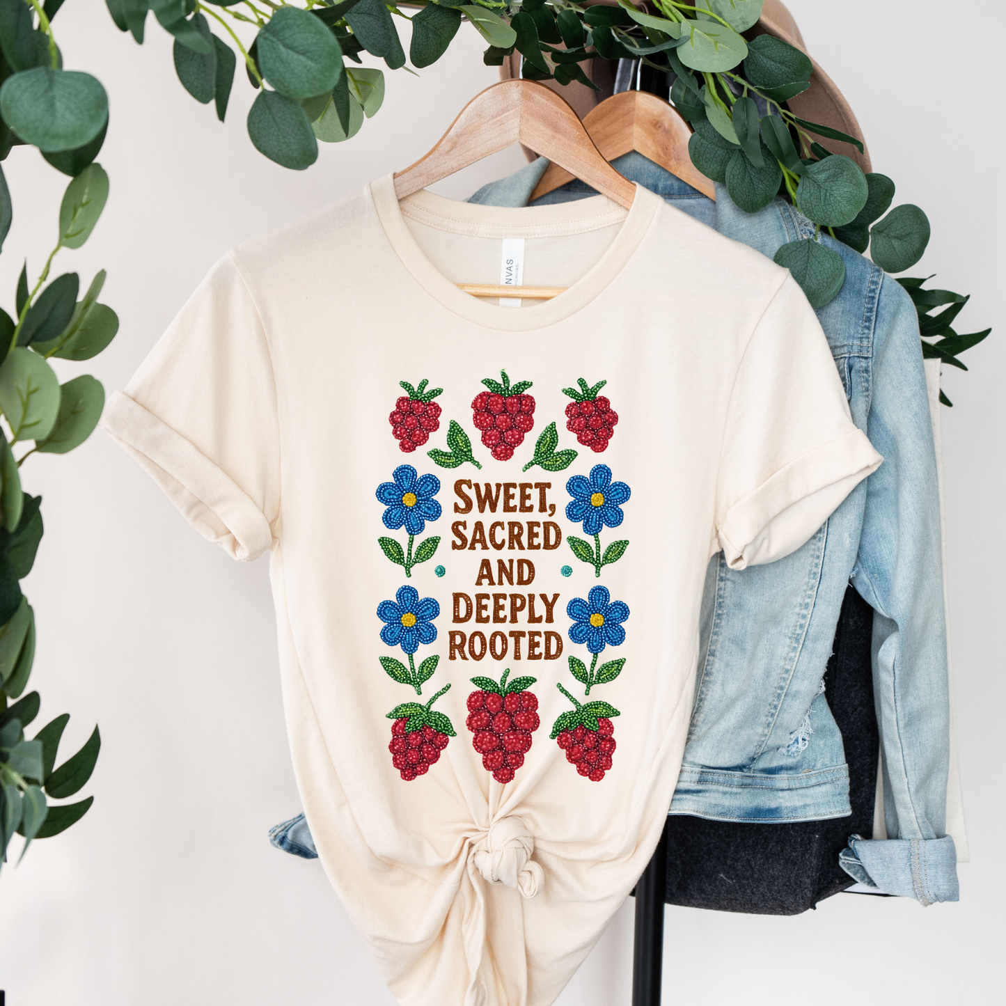 Beige t-shirt with floral design and text on a hanger with greenery and denim jacket in the background. sweet sacred deeply rooted shirt, beaded floral berry tee, indigenous beaded design shirt, sacred roots indigenous apparel, floral berry cultural t-shirt, original indigenous graphic tee, beaded indigenous shirt, deeply rooted heritage tee, unisex cultural pride shirt, sahdeh spruce indigenous design, berry beadwork t-shirt, ancestral connection apparel