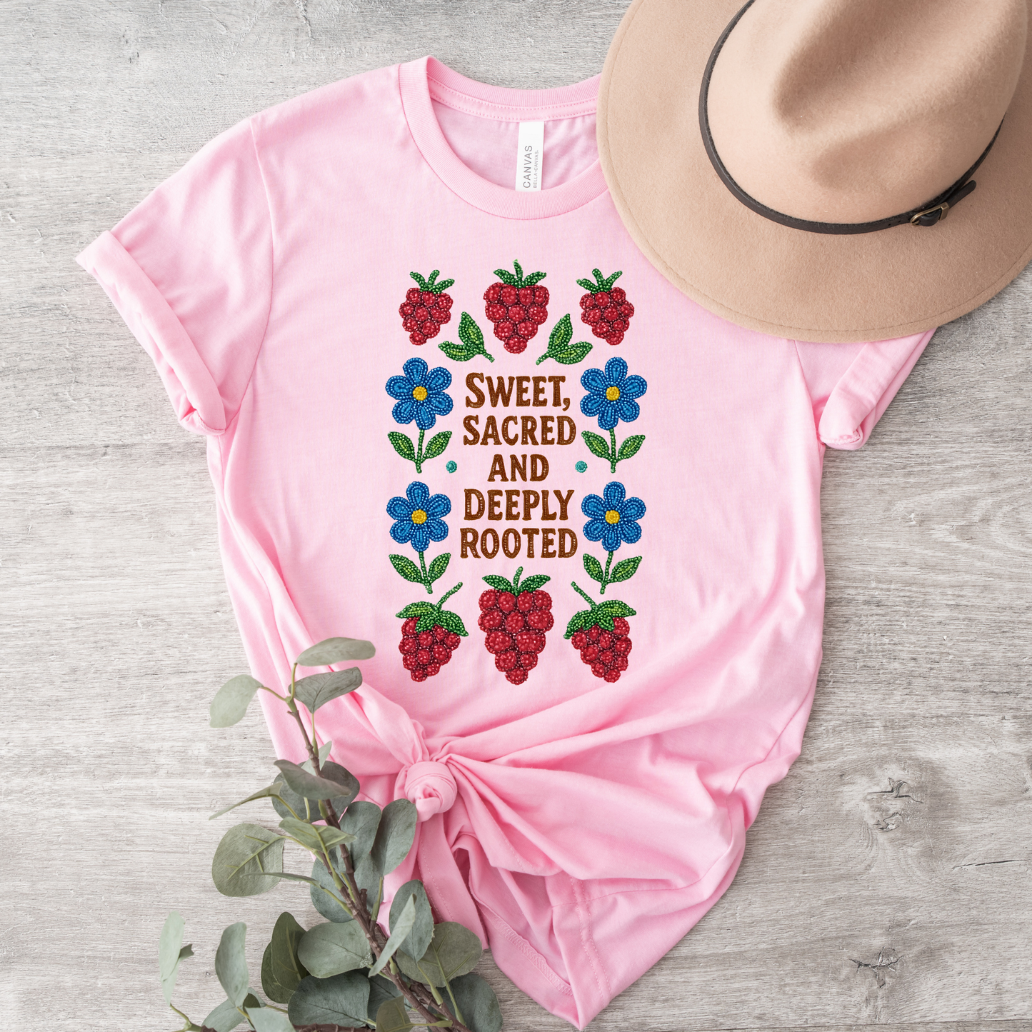 Pink t-shirt with floral design and text, accompanied by a hat and plant on a wooden surface. sweet sacred deeply rooted shirt, beaded floral berry tee, indigenous beaded design shirt, sacred roots indigenous apparel, floral berry cultural t-shirt, original indigenous graphic tee, beaded indigenous shirt, deeply rooted heritage tee, unisex cultural pride shirt, sahdeh spruce indigenous design, berry beadwork t-shirt, ancestral connection apparel