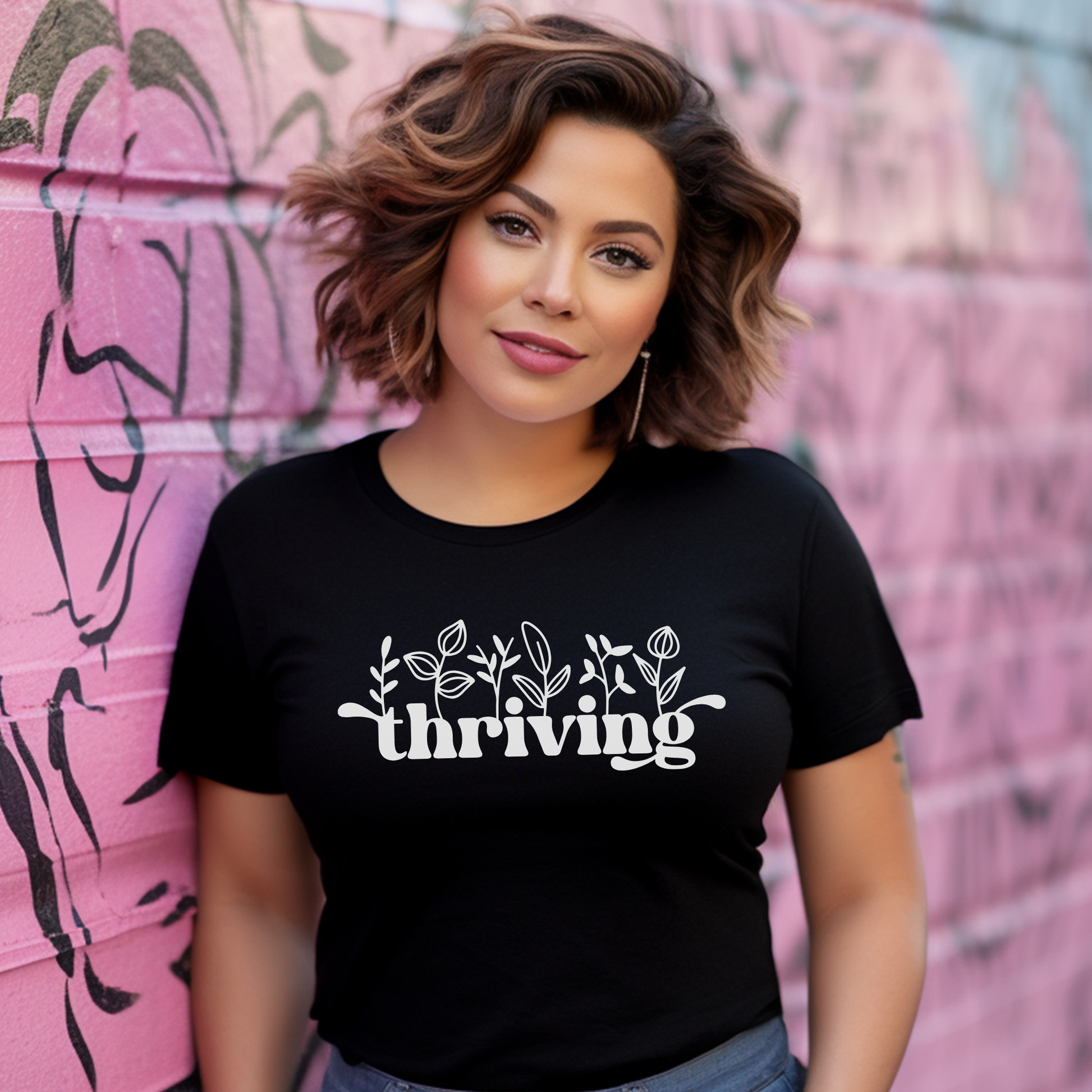 A woman with wavy brown hair wearing a black t-shirt with a white graphic that reads 'thriving' under small leaf and flower illustrations, thriving shirt, floral graphic tee, positive affirmation shirt, inspirational shirt, motivational tee, unisex graphic tee, botanical shirt, empowering apparel, everyday wear shirt, confidence shirt, uplifting gift, statement shirt, positive mindset tee, floral typography shirt, casual inspirational tee